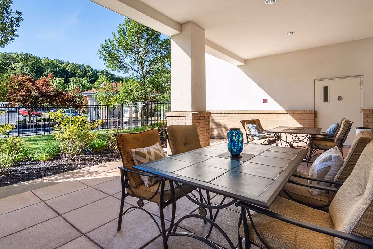 Covered outdoor patio area with metal tables and cushioned chairs, surrounded by greenery and trees with a parking lot visible in the background under a clear blue sky.