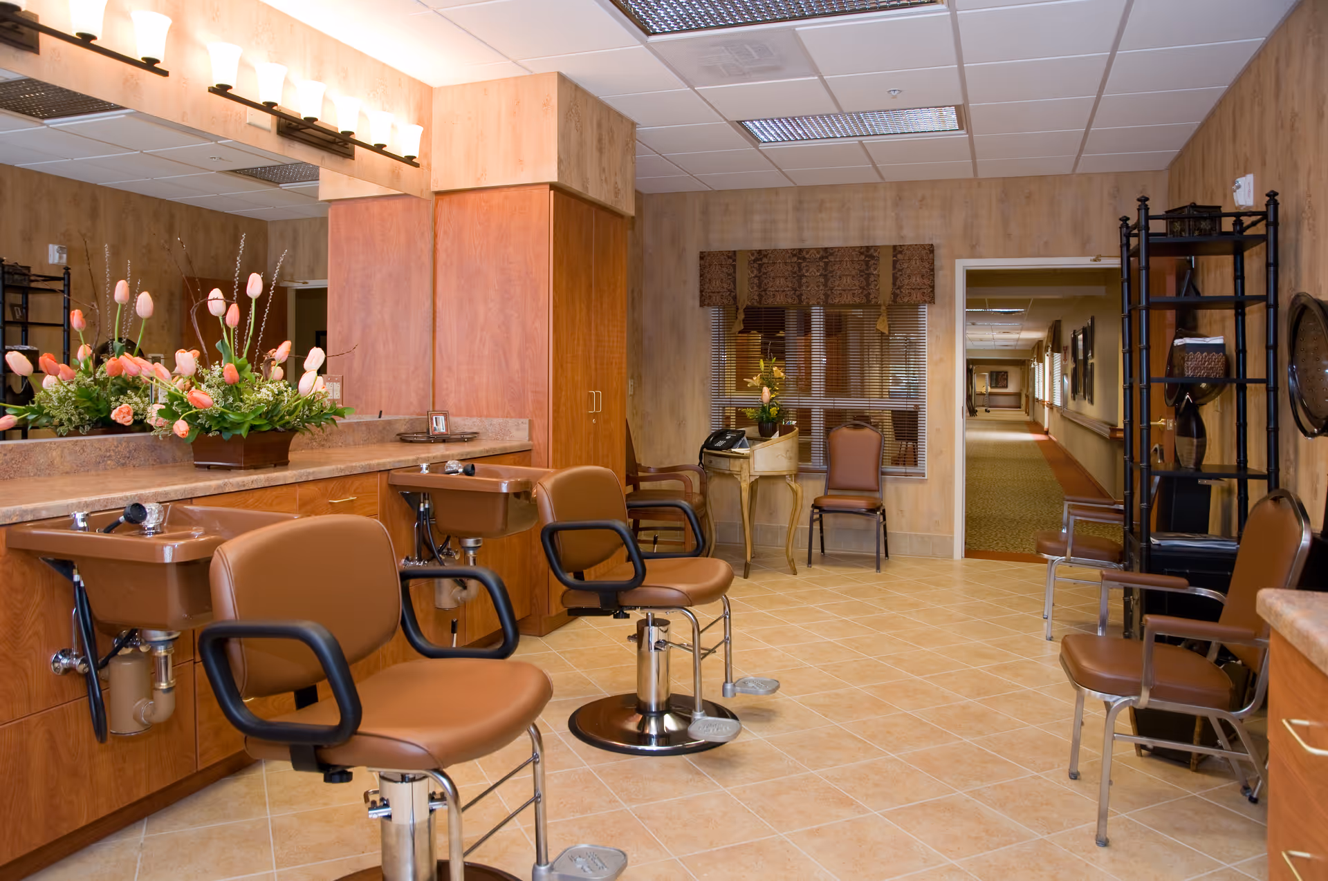A well-lit salon in a senior living facility with brown styling chairs, wash basins, a large mirror, and a floral arrangement.