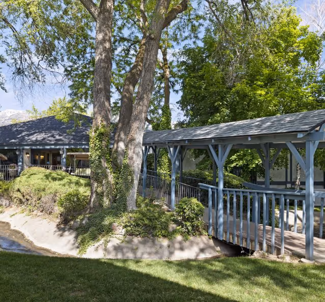 Outdoor scene at Cove Point Retirement featuring a wooden covered bridge over a small stream, surrounded by green trees and bushes, with a building visible in the background under a clear sky.
