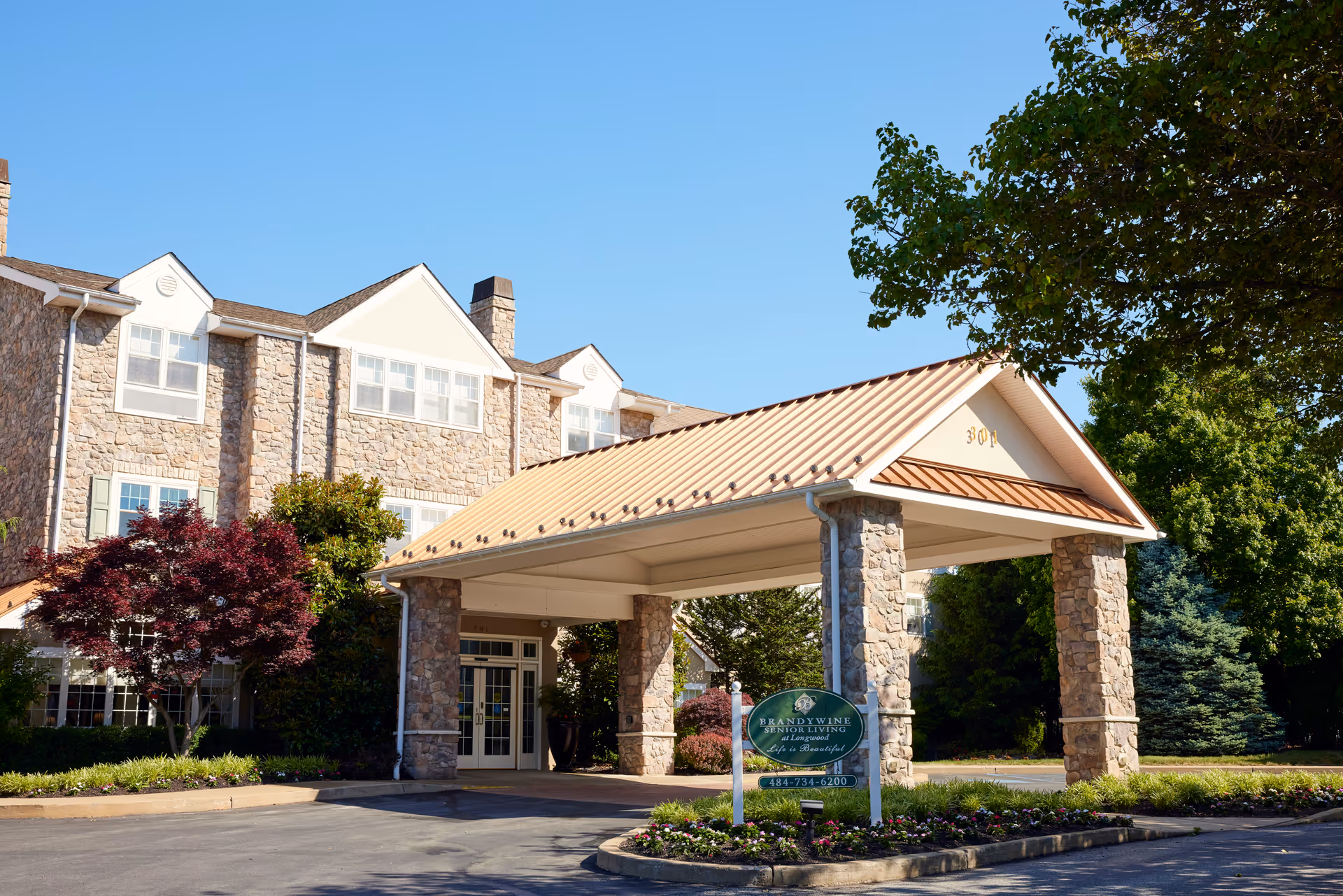 Exterior view of Brandywine Longwood by Monarch senior living facility showing a stone building with multiple windows, a covered entrance with stone pillars, and a sign with the facility name and phone number. Trees and landscaping surround the entrance under a clear blue sky.