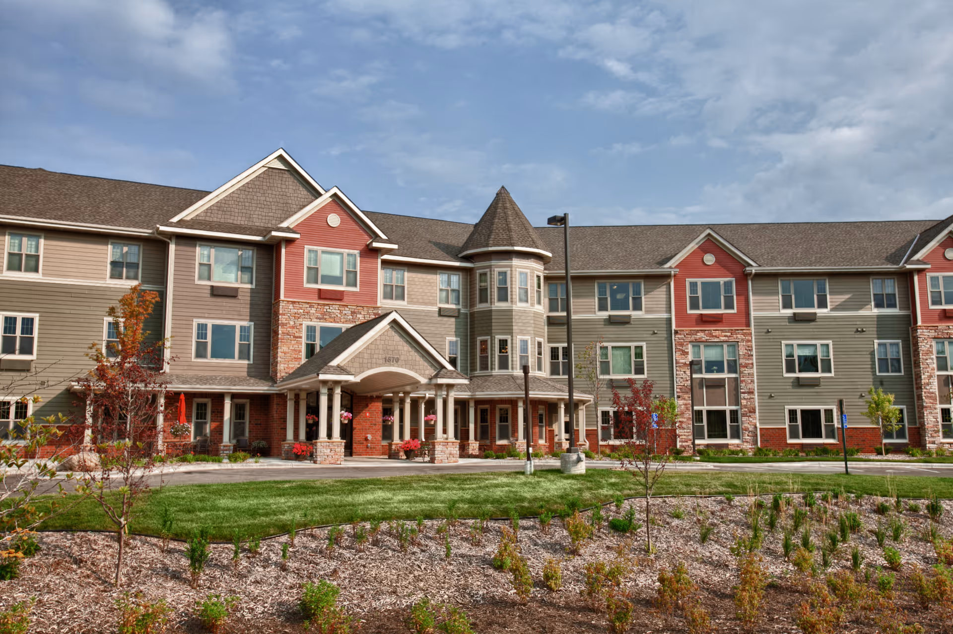 Front exterior of a three-story senior living building with a covered entrance, turret, and landscaped lawn.