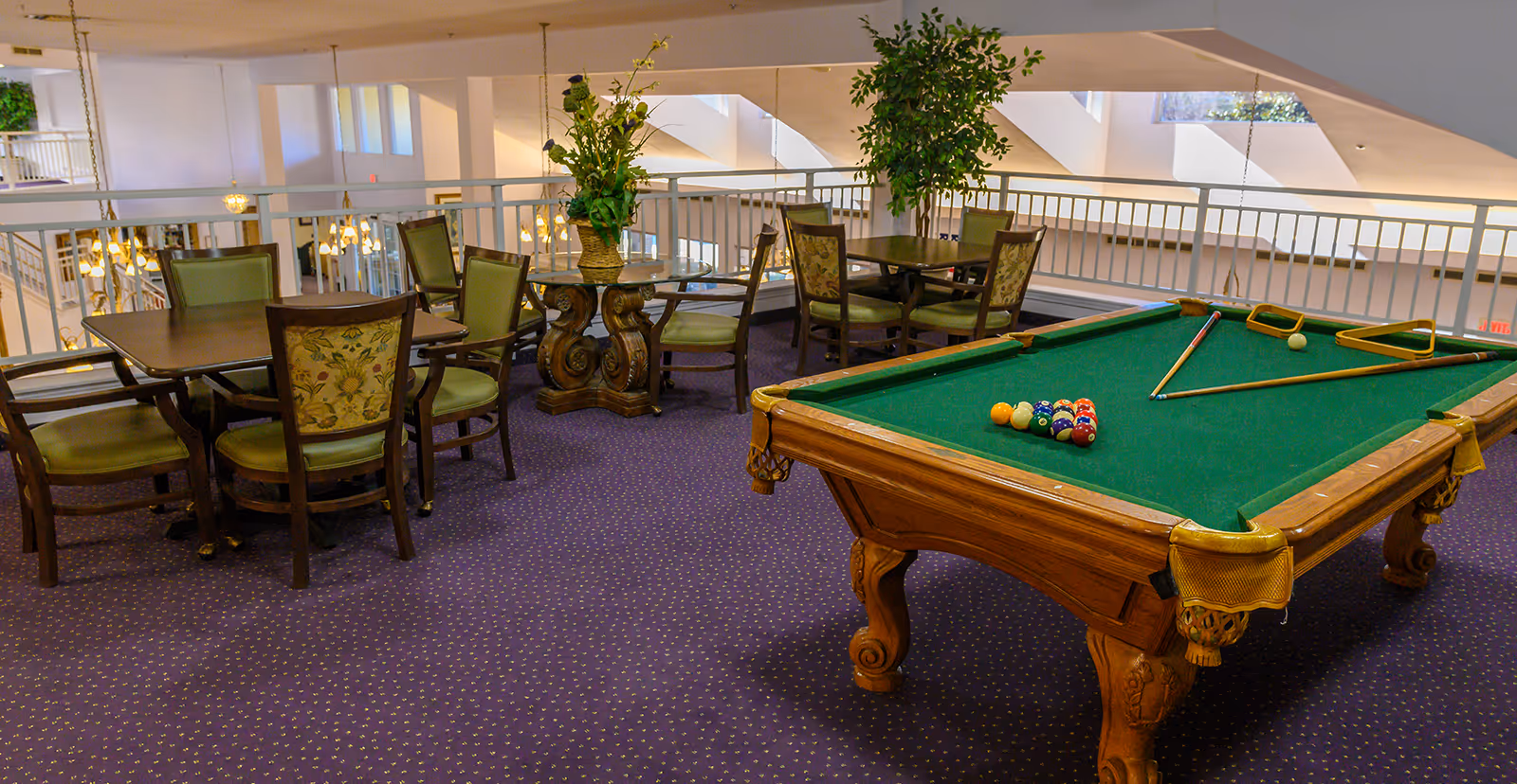 Interior view of a senior living facility's recreational area featuring a pool table with balls and cues set up, surrounded by several tables and chairs with floral upholstery and green cushions. The space has purple carpet, decorative plants, and overlooks a lower level with hanging lights and large windows allowing natural light.