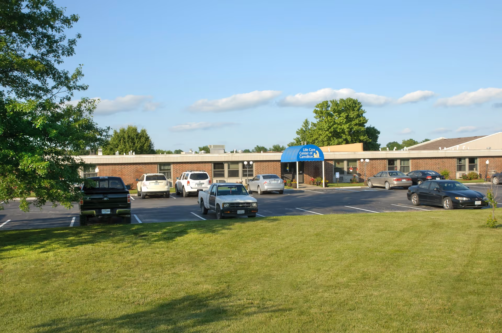 Front exterior of a single-story brick Life Care Center of Carrollton with a parking lot, several cars, and a grassy lawn.