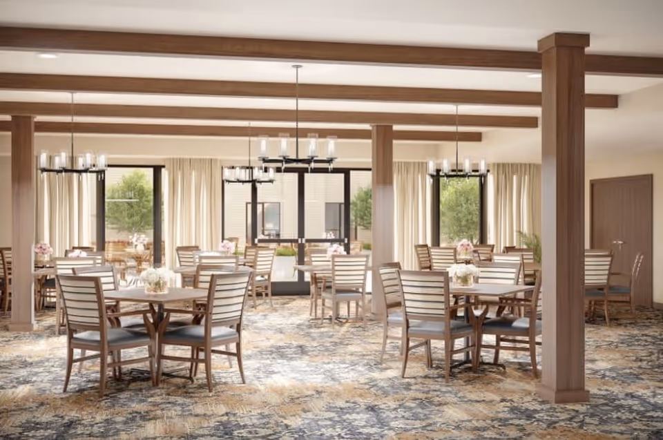 A spacious dining room with multiple wooden tables and chairs arranged neatly. The room features large windows with cream-colored curtains, wooden beams on the ceiling, and modern chandeliers hanging above the tables. The carpet has a patterned design in shades of blue, beige, and gray. There are flower arrangements on each table, and the room has a warm, inviting atmosphere.