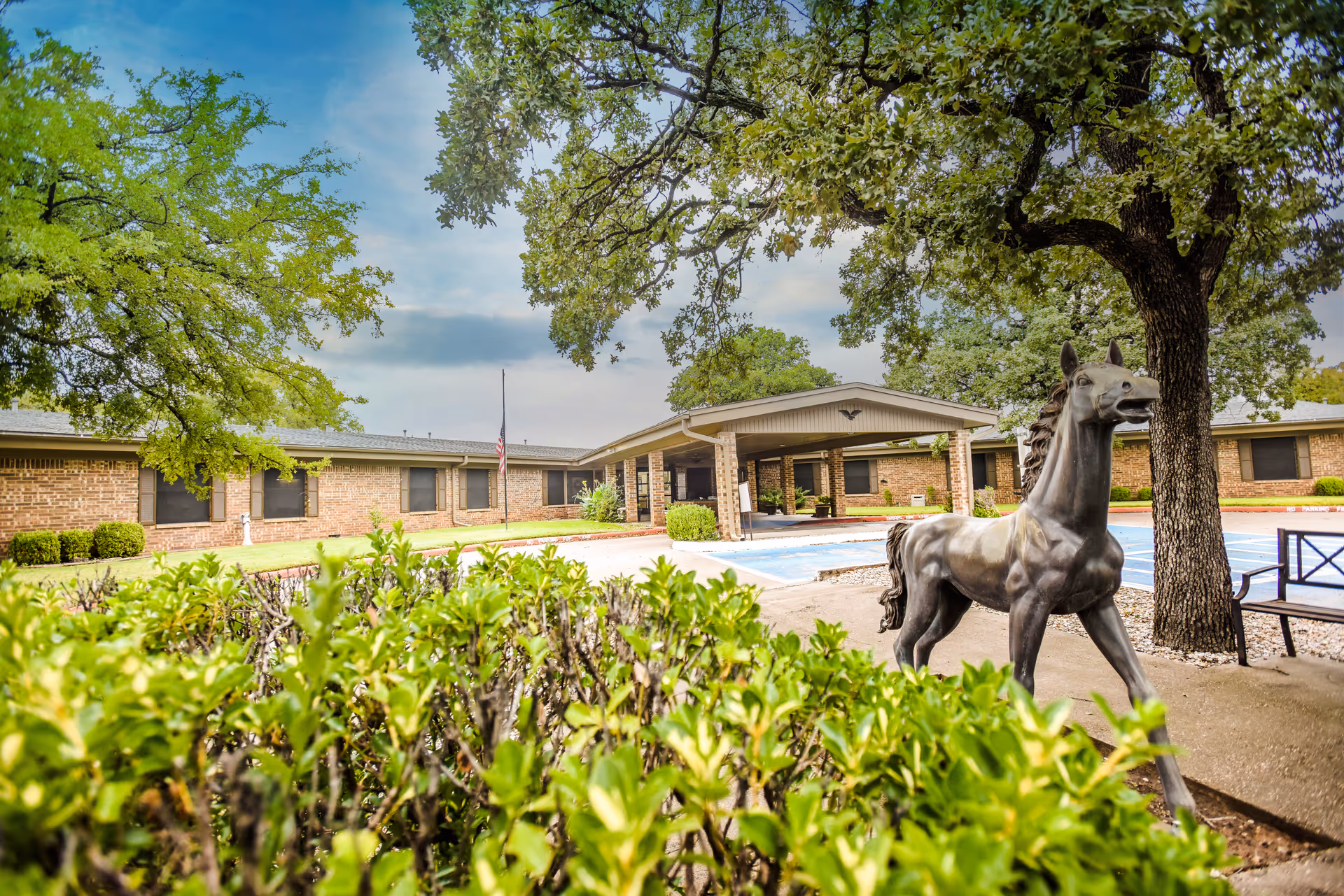 Exterior view of Graham Oaks Care Center showing a single-story brick building with multiple windows, surrounded by green trees and bushes. In the foreground, there is a bronze statue of a horse near a tree and a bench. The sky is partly cloudy.