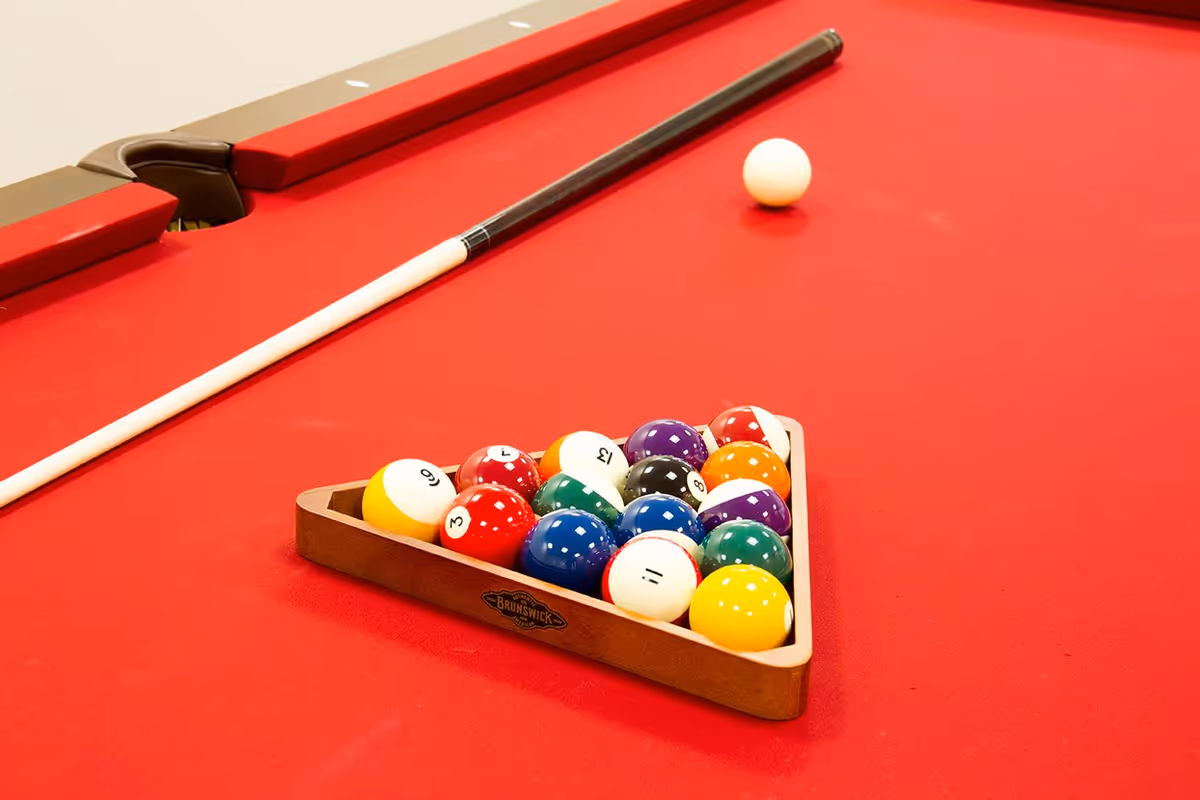 A red pool table with a wooden triangle rack holding a full set of colorful billiard balls and a white cue ball placed outside the rack. A pool cue rests on the edge of the table.