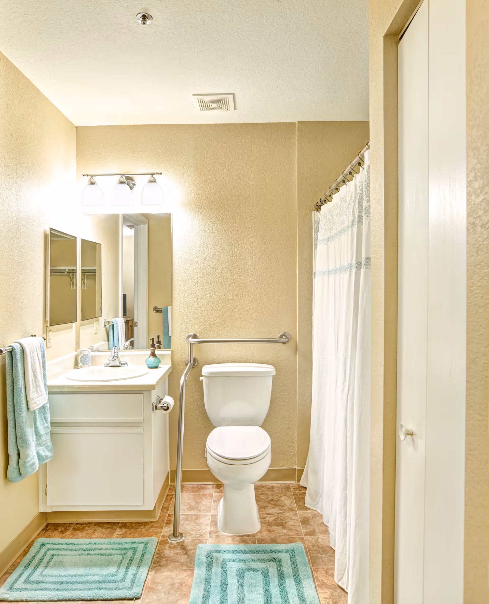 A clean bathroom with beige walls and tiled floor featuring a white toilet with a support rail, a white vanity with a sink, a mirror above it, and a three-light fixture. There are two teal bath mats on the floor, teal towels hanging on a towel rack, and a white shower curtain with a subtle blue pattern.