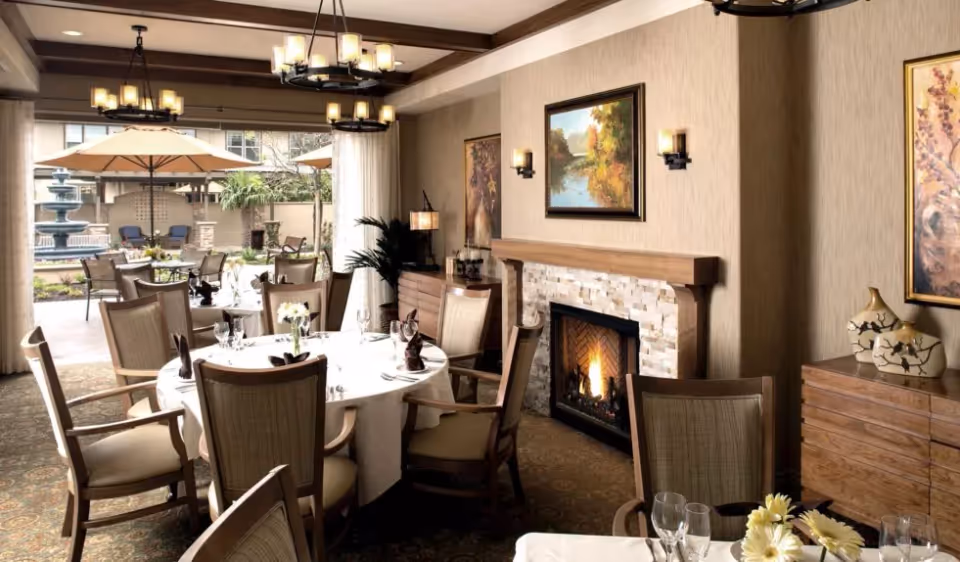 A cozy dining room with a round table set for a meal, surrounded by wooden chairs with cushioned seats. The room features a lit fireplace with a stone surround, framed paintings on the walls, and warm lighting from wall sconces and chandeliers. Large windows and an open doorway reveal an outdoor patio area with tables, chairs, umbrellas, and a fountain.