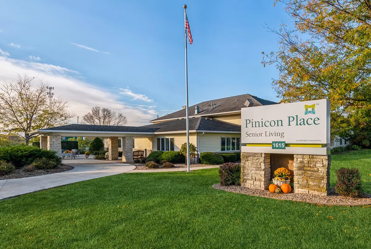Exterior view of Pinicon Place Senior Living facility with a large sign displaying the name and address, a flagpole with an American flag, green lawn, trees, and a covered entrance with benches and shrubs.