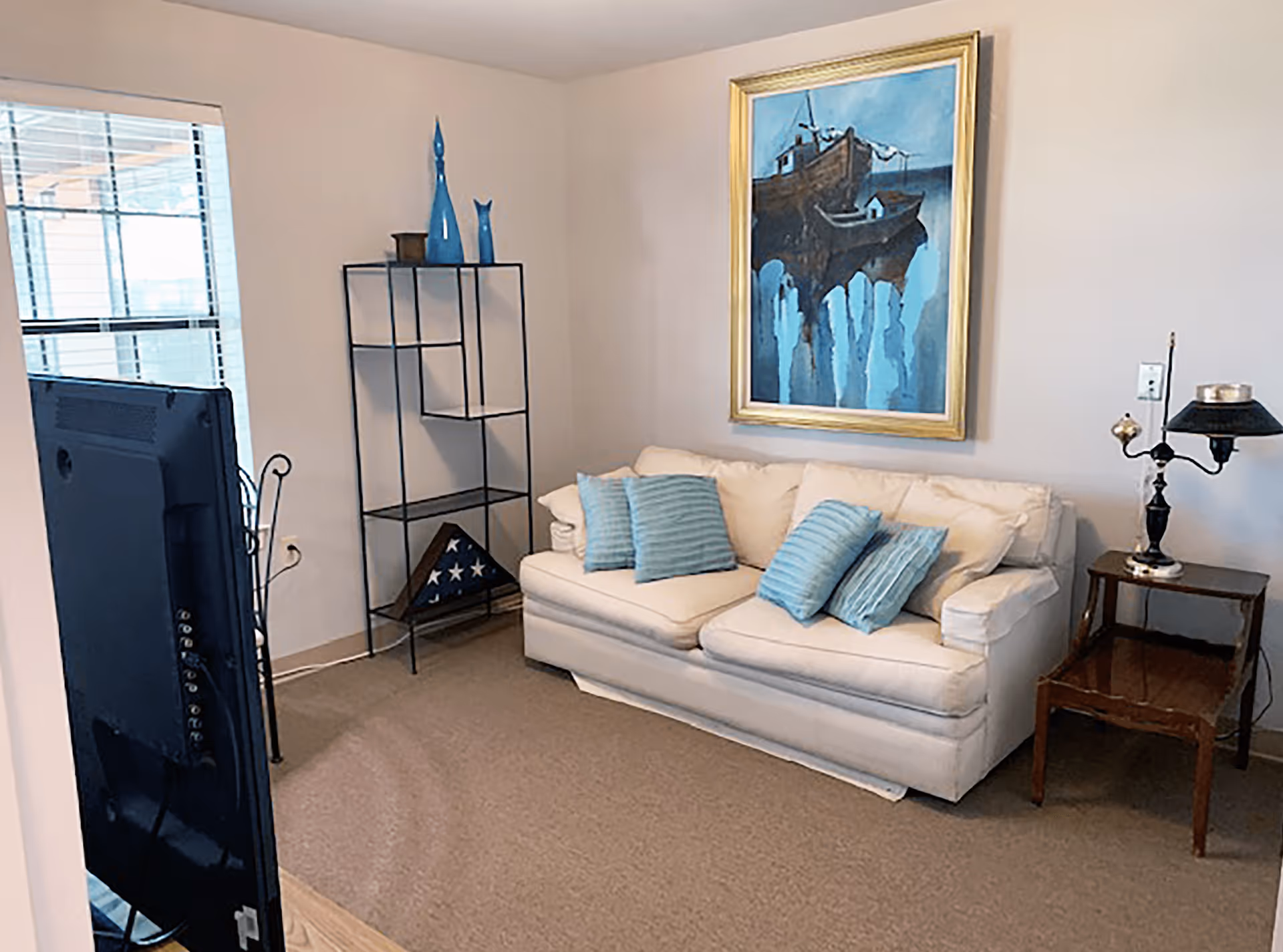 A cozy living room with a white sofa adorned with four blue striped pillows. Above the sofa hangs a framed painting of boats on water in shades of blue. To the right of the sofa is a wooden side table with a vintage-style lamp. To the left, there is a black metal shelving unit with decorative blue vases and a folded American flag displayed on the bottom shelf. A large window with blinds is visible on the left wall, and a flat-screen TV is partially visible in the foreground.