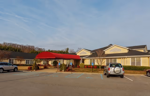 Front exterior of a single-story yellow senior living facility with a red entrance canopy and adjacent parking lot.