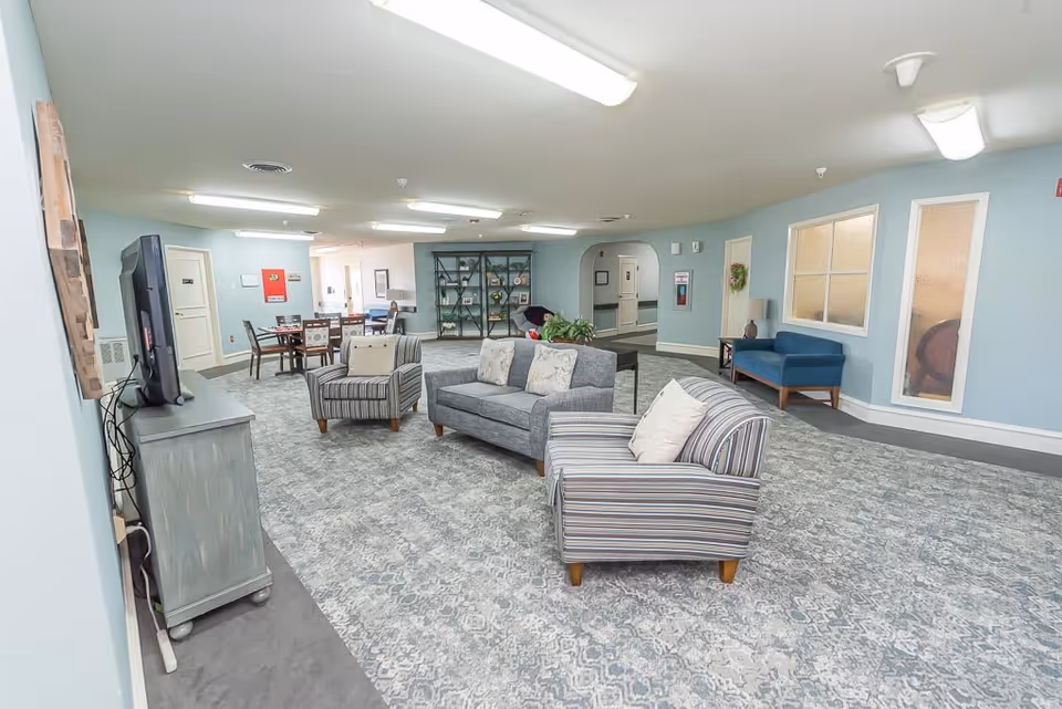 A spacious senior living facility common area with light blue walls and patterned carpet. The room features a gray sofa with two pillows, two striped armchairs, a blue loveseat, a TV on a gray cabinet, and a dining table with chairs in the background. There is a bookshelf with decorative items and plants, and several doors and windows along the walls.