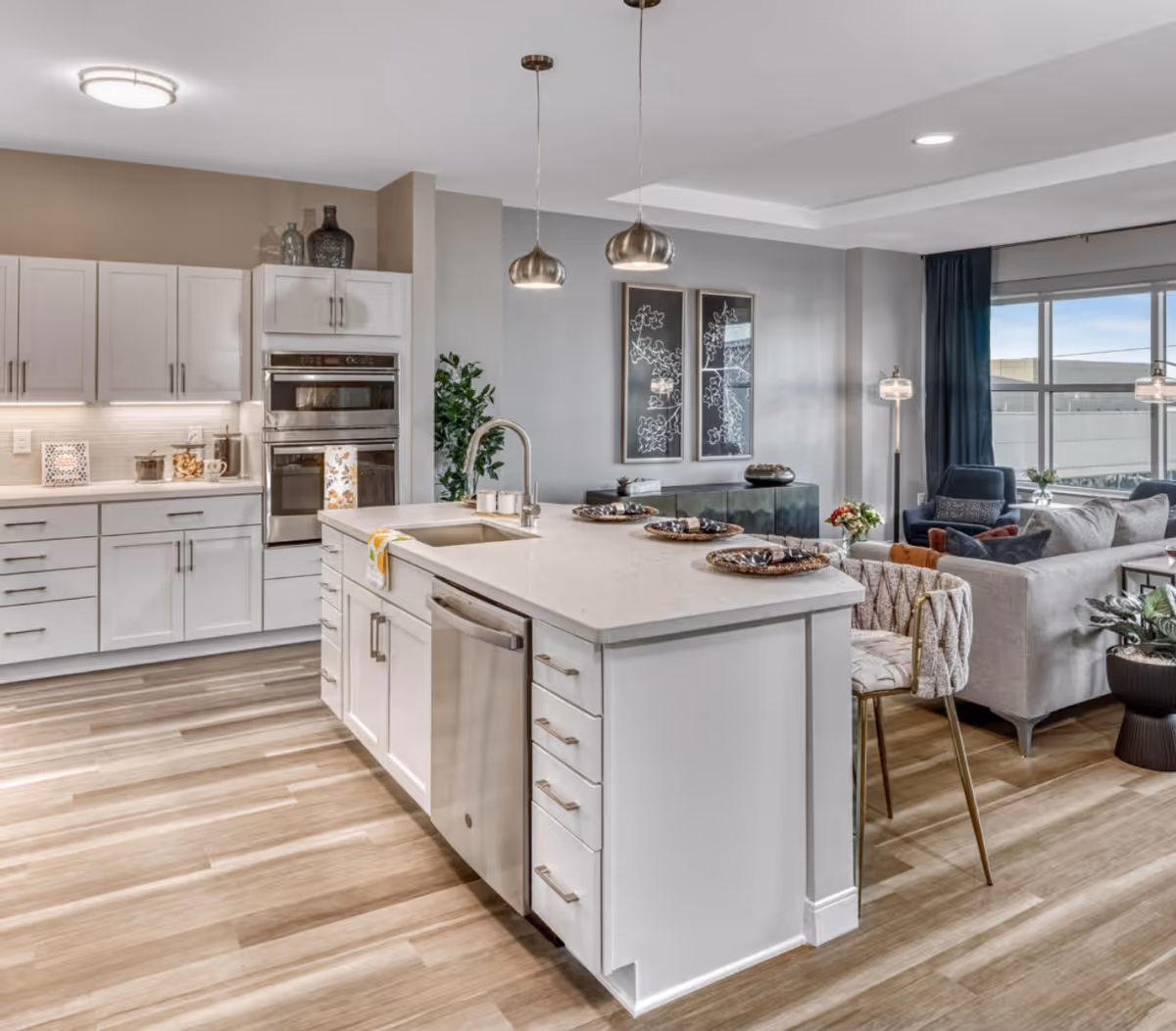 Bright open-plan kitchen with a large white island, stainless appliances, pendant lights, and a living area visible in the background.