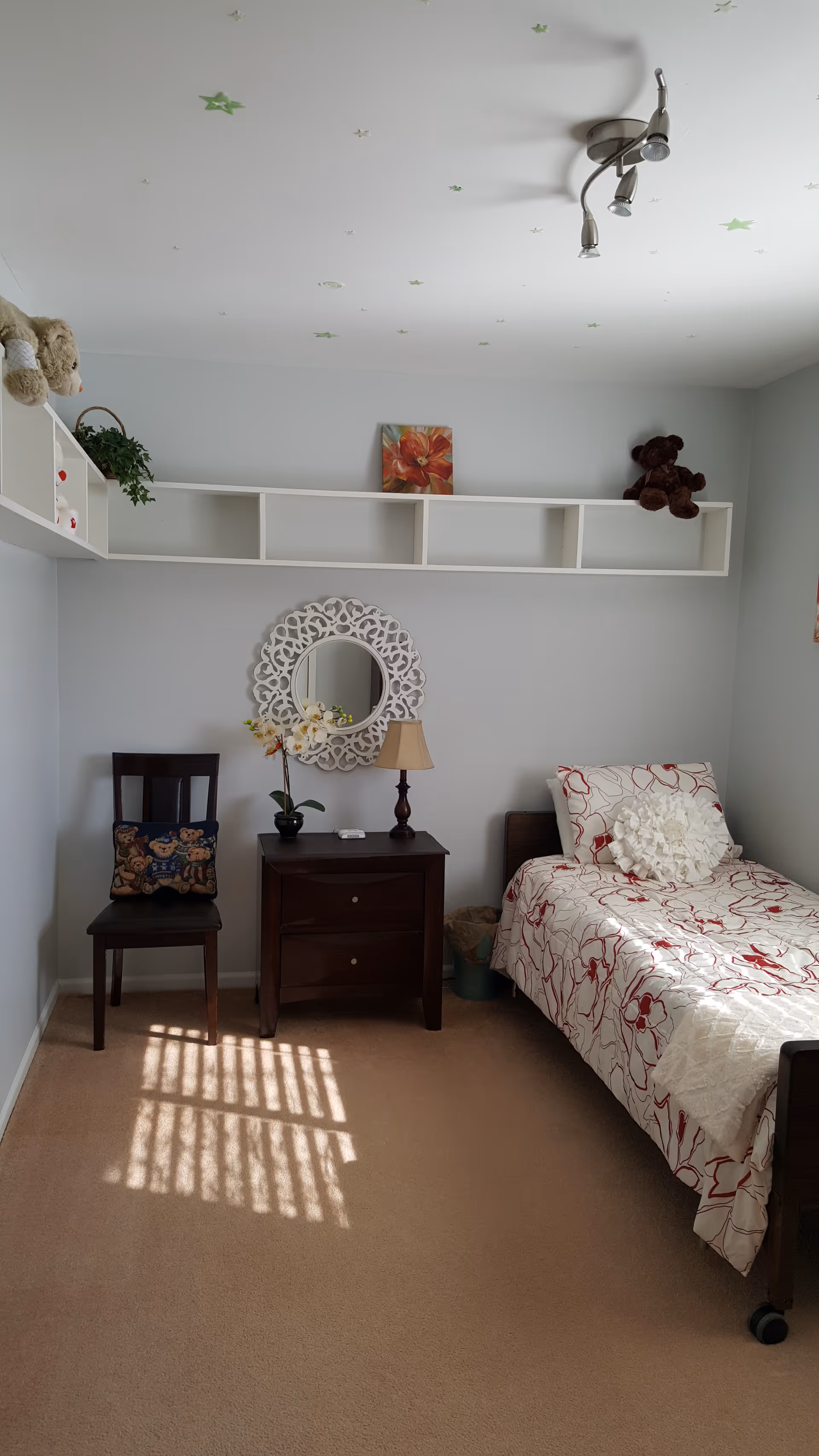 A small, cozy bedroom with a single bed covered in a white and red floral bedspread. Next to the bed is a dark wooden nightstand with a lamp and a small plant. A dark wooden chair with a decorative pillow is placed beside the nightstand. Above the nightstand is a decorative round mirror with a white ornate frame. A long white shelf runs along the wall near the ceiling, holding a few decorative items including a teddy bear and a small painting. The ceiling has small star stickers and a modern light fixture. Sunlight casts a shadow pattern on the beige carpeted floor.