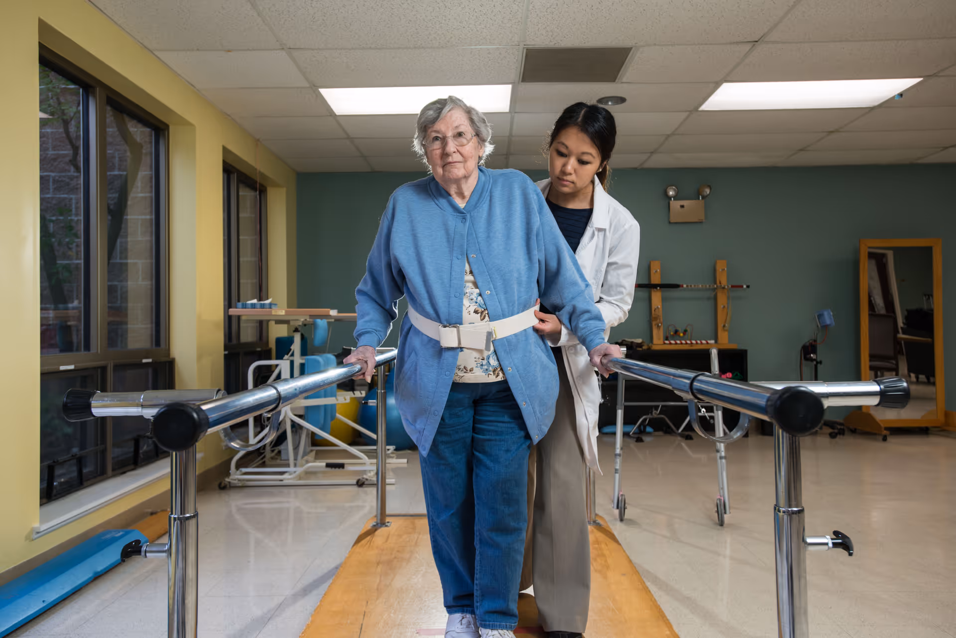 An elderly woman wearing a blue cardigan and jeans is walking with the support of parallel bars in a rehabilitation room. A healthcare professional in a white coat is assisting her by holding a gait belt around her waist. The room has large windows, exercise equipment, and a mirror in the background.