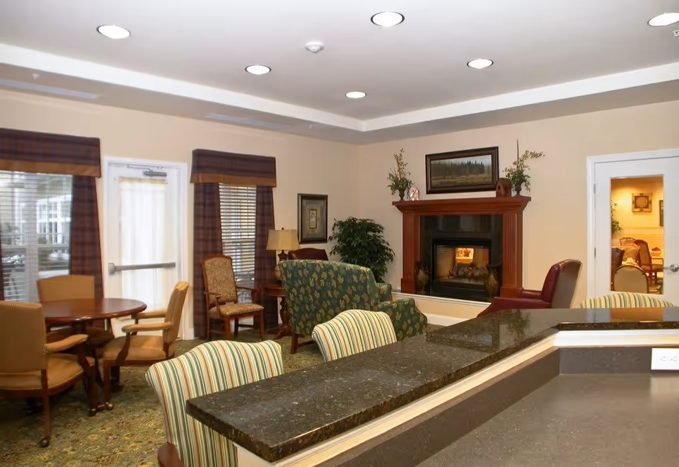 A cozy living room area in a senior living facility featuring a fireplace with a wooden mantel decorated with plants and a framed picture above it. The room has several chairs including striped upholstered chairs, a green patterned armchair, and brown leather chairs. There is a round wooden table with chairs near windows covered with plaid valances, and a countertop with a dark granite surface in the foreground.