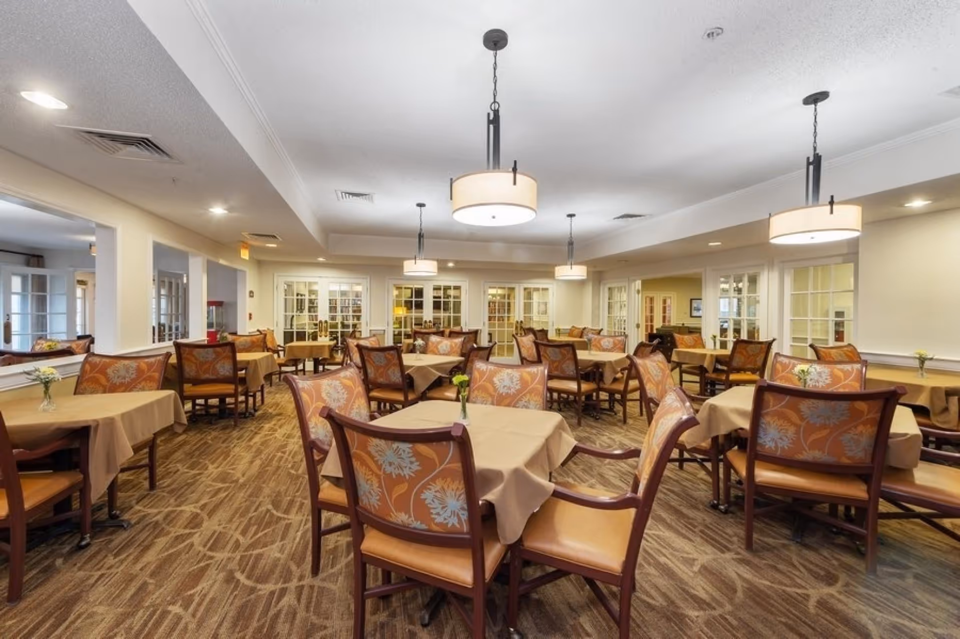 Bright dining room with multiple cloth-covered tables and patterned chairs under hanging pendant lights.