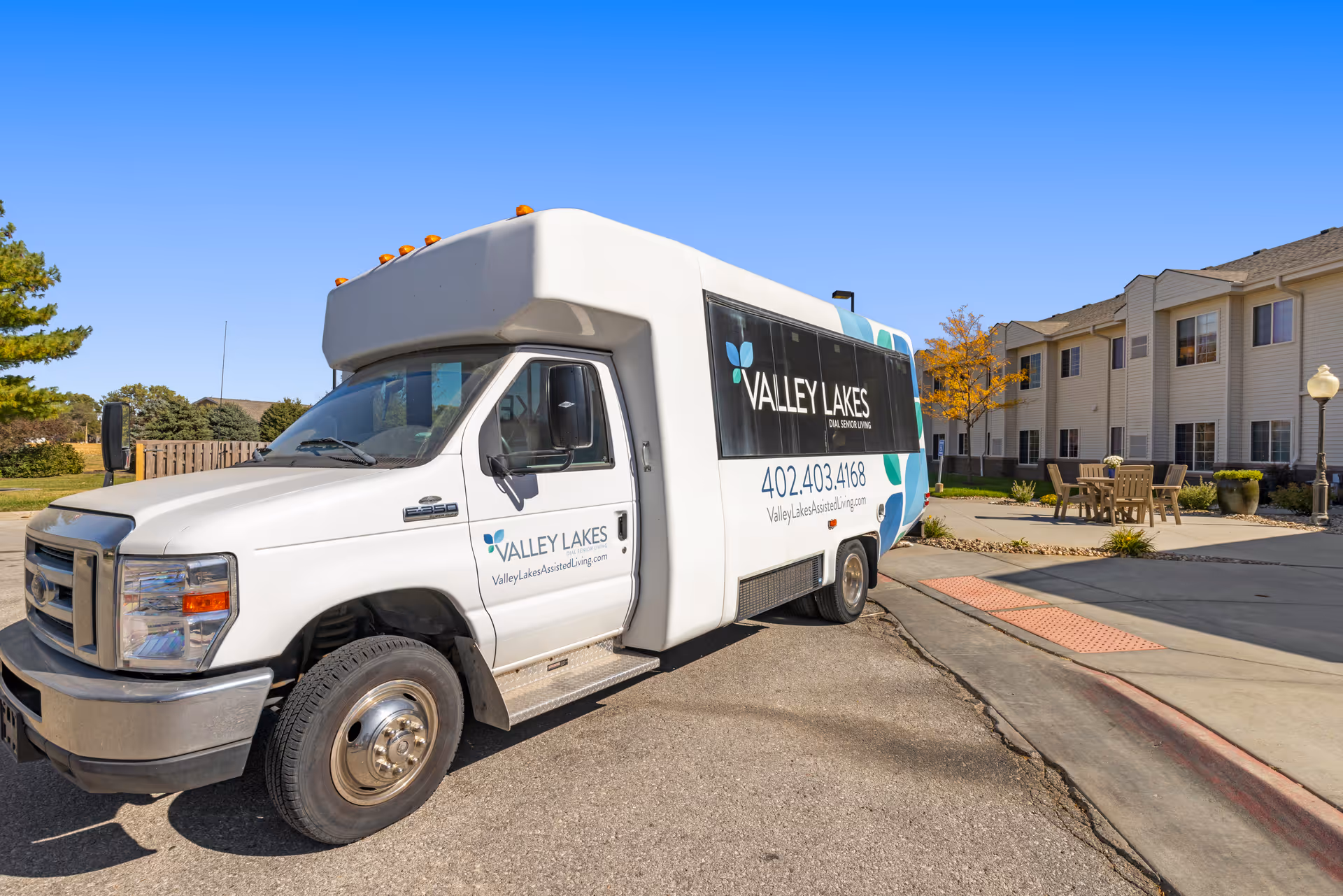 A white shuttle bus parked outside a senior living facility named Valley Lakes Assisted Living. The bus has the facility's name, logo, phone number, and website on its side. In the background, there is a two-story building with multiple windows, outdoor seating with tables and chairs, a tree with autumn-colored leaves, and a clear blue sky.