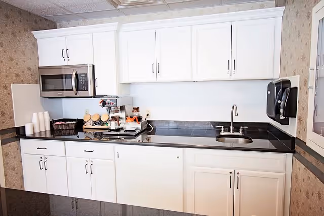 A clean kitchen area with white cabinets, a black countertop, a microwave mounted above the counter, a coffee maker with cups and condiments, a small sink with a faucet, and a wall-mounted soap dispenser.