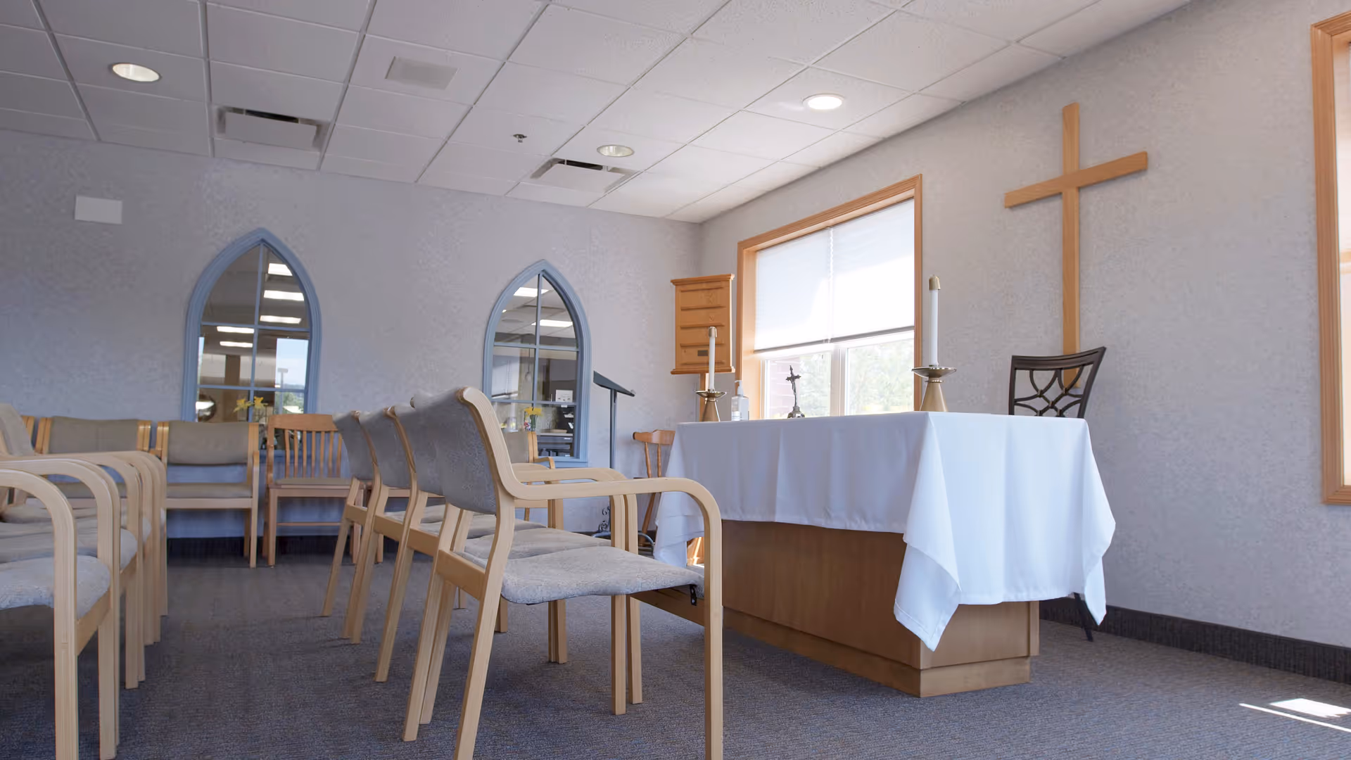 Interior view of a small chapel or prayer room with rows of wooden chairs with gray cushions facing an altar covered with a white cloth. On the altar are two candles and a small cross. A large wooden cross is mounted on the wall behind the altar. The room has light-colored walls, two arched mirrors, and a window with a white blind letting in natural light.