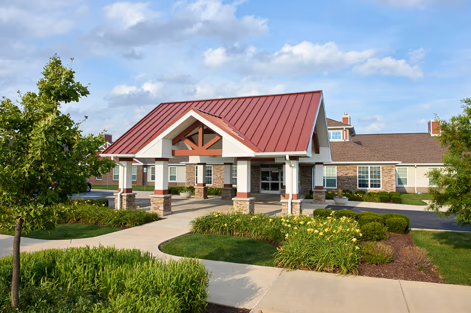 Exterior view of a senior living facility entrance with a red metal roof canopy supported by white columns with stone bases. The building has beige siding and stone accents, surrounded by well-maintained landscaping including green bushes, yellow flowers, and trees under a partly cloudy sky.