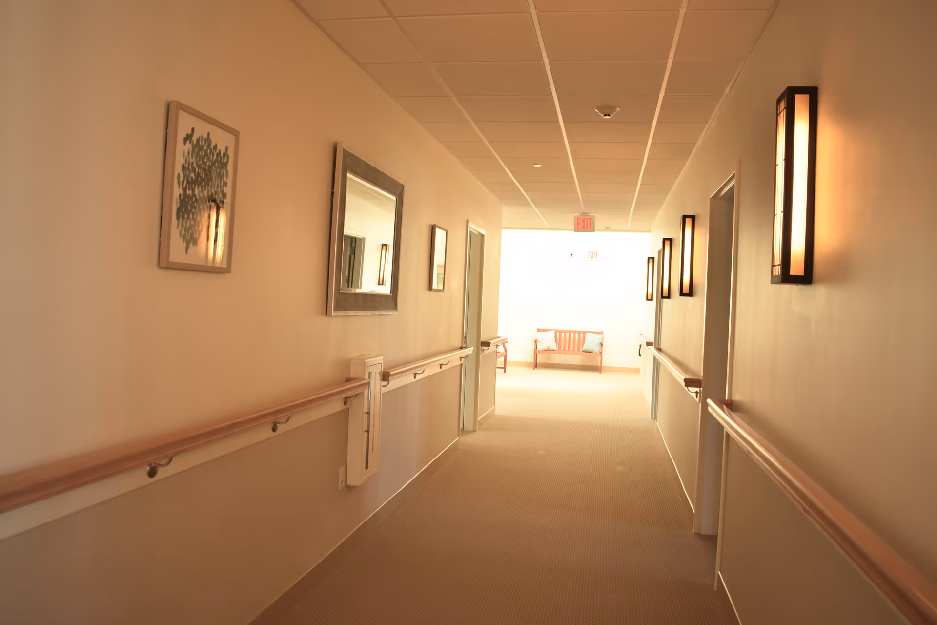 A well-lit hallway in a senior living facility with handrails on both sides, wall-mounted lights, framed artwork, and a bench with pillows at the far end near an exit sign.