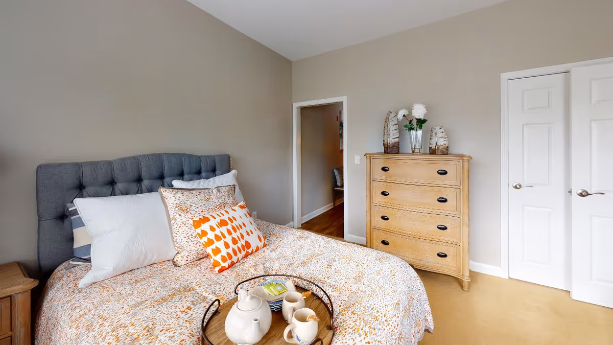 A bedroom with a gray tufted headboard bed dressed in white and orange patterned bedding with multiple pillows. A wooden nightstand is beside the bed. Across from the bed is a wooden dresser with decorative items including a vase with white flowers and ornamental feathers. The room has beige walls, a beige carpet, and white double closet doors. An open doorway leads to another room with wooden flooring.