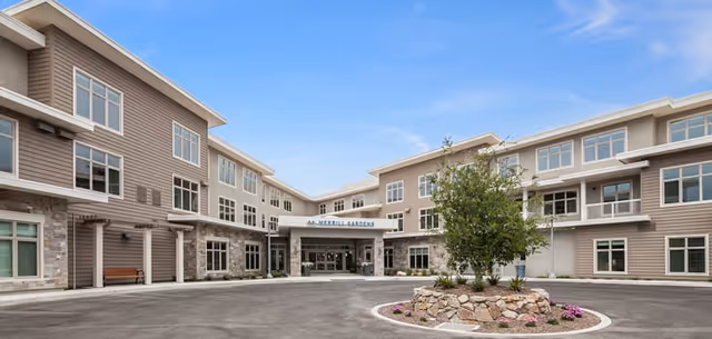 Exterior view of Merrill Gardens at Monterey, showing a three-story building with multiple windows, a central entrance with a covered driveway, and a landscaped circular island with a tree and flowers in front.