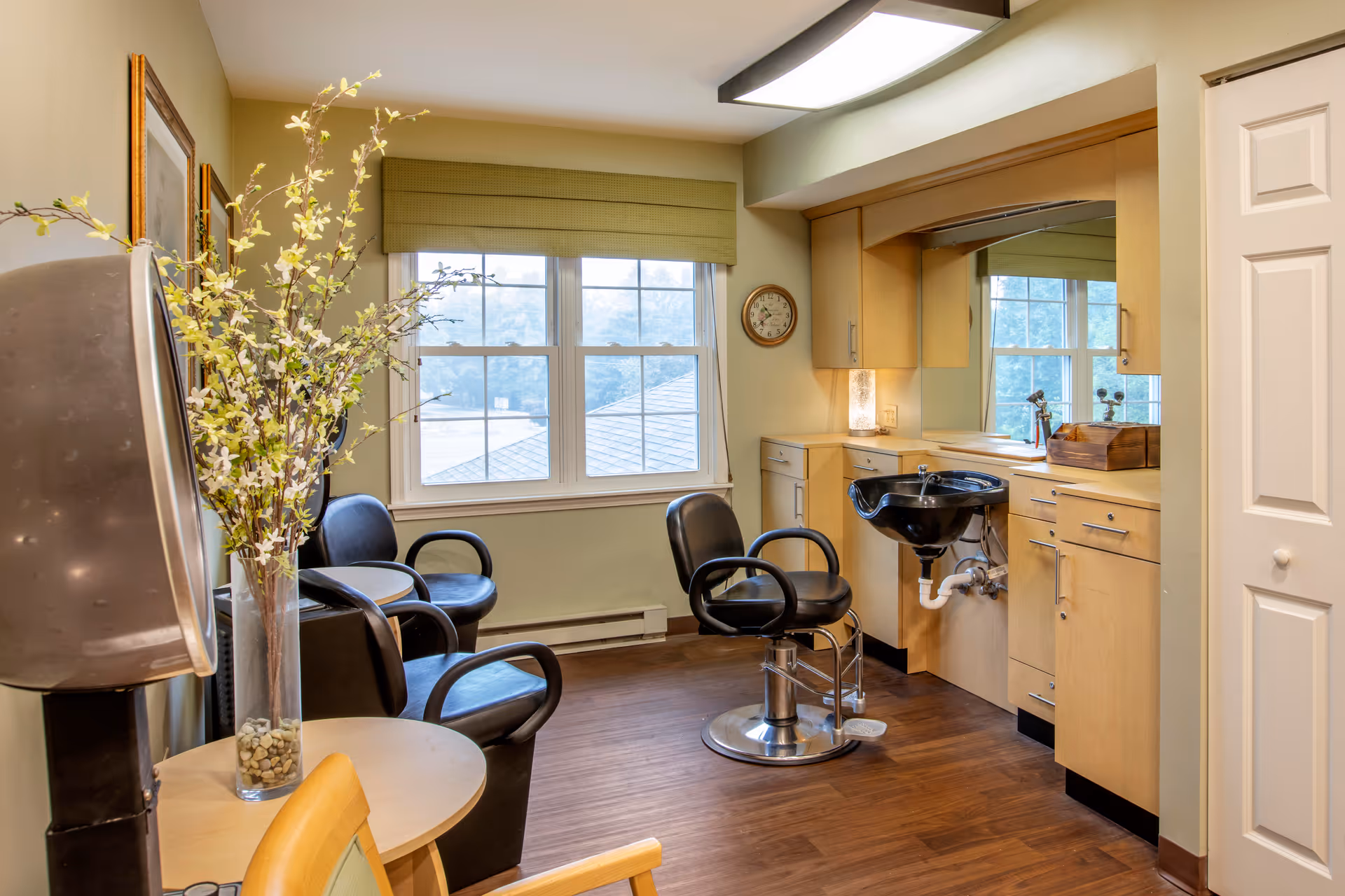 A bright and clean hair salon room with a black salon chair in front of a black sink, light wood cabinetry with a large mirror, and a window with a green valance letting in natural light. There are additional black chairs along the wall and a tall vase with white flowers on a small round table.