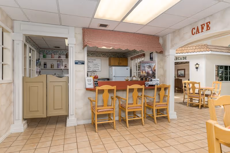 Interior view of a cozy cafe area in a senior living facility named Quail Ridge, featuring a counter with four wooden chairs with checkered cushions, a small kitchen area behind the counter with a refrigerator and shelves, and additional seating with tables and chairs in the background under a sign that reads 'CAFE'.