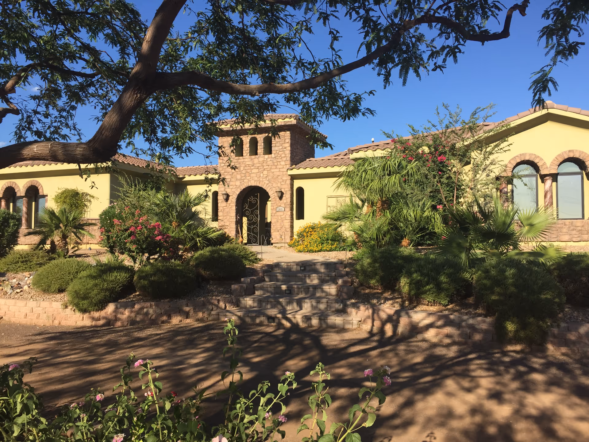 Exterior view of a single-story building with a stone entrance tower and yellow walls, surrounded by lush greenery, bushes, and trees under a clear blue sky.