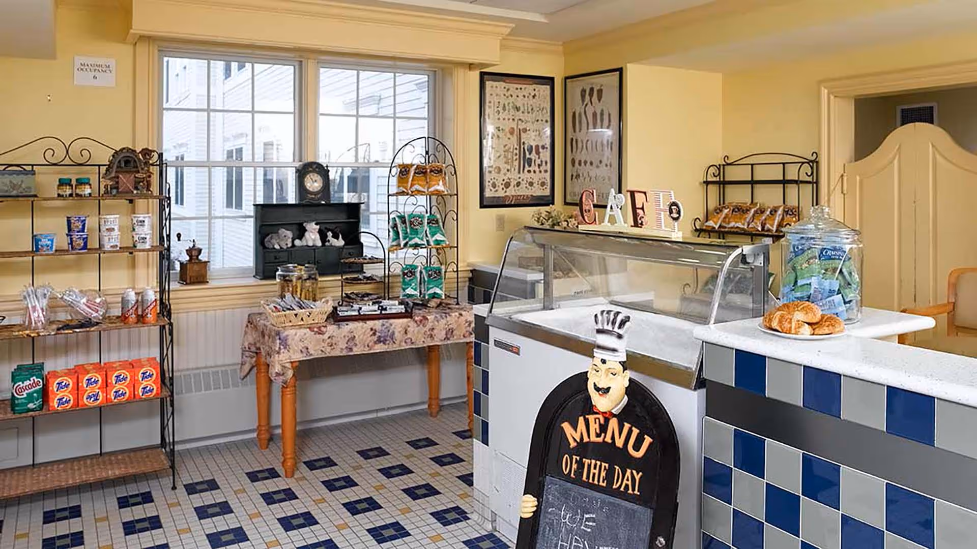A small cafe area inside a senior living facility with shelves stocked with snacks and household items, a table with more snacks, a glass display counter, and a signboard shaped like a chef that reads 'Menu of the Day'. The room has yellow walls, a window, and a tiled floor with a blue and beige pattern.