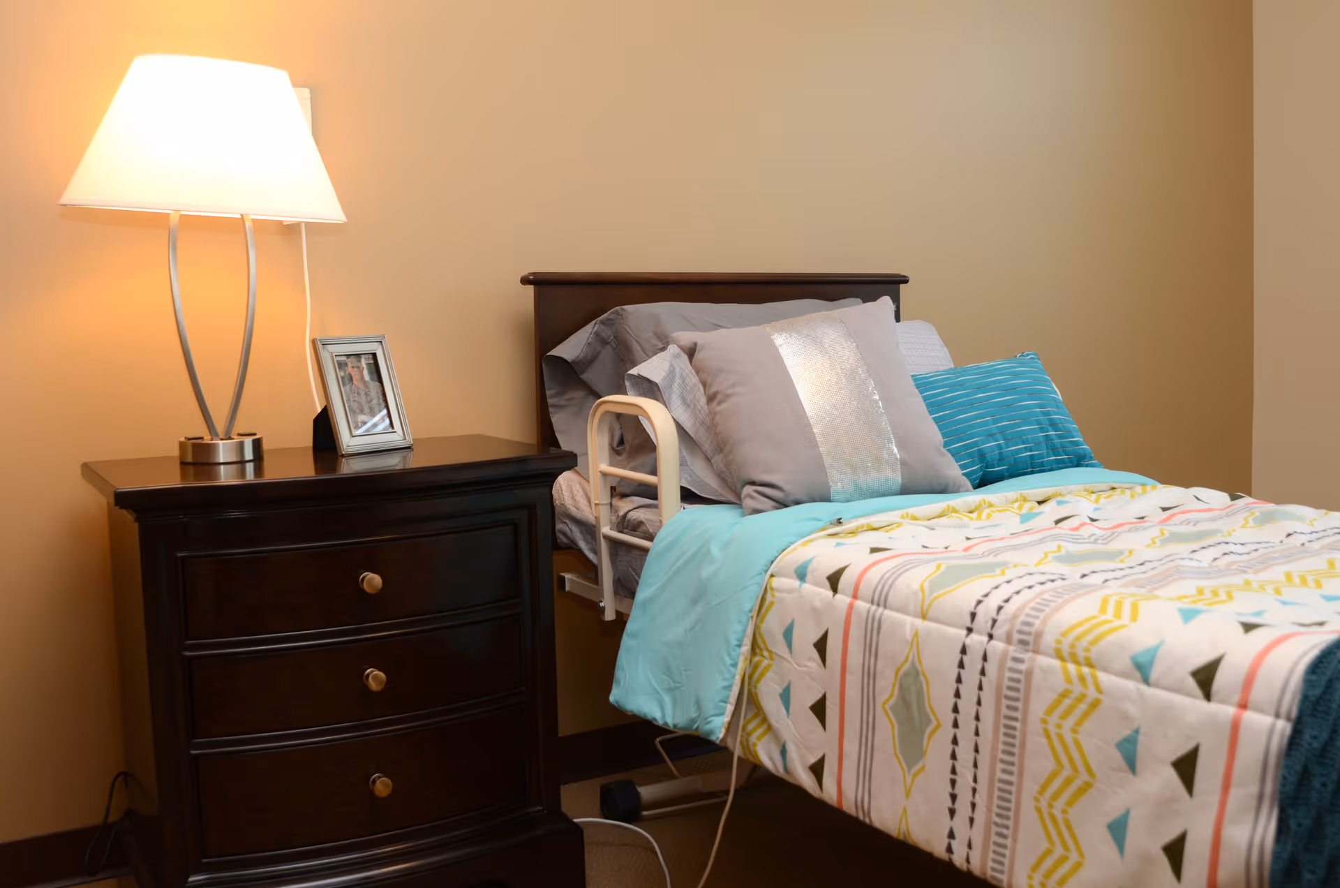 A neatly made single bed with multiple pillows and a colorful patterned blanket in a room with beige walls. Next to the bed is a dark wooden nightstand with three drawers, a modern lamp, and a framed photo on top.