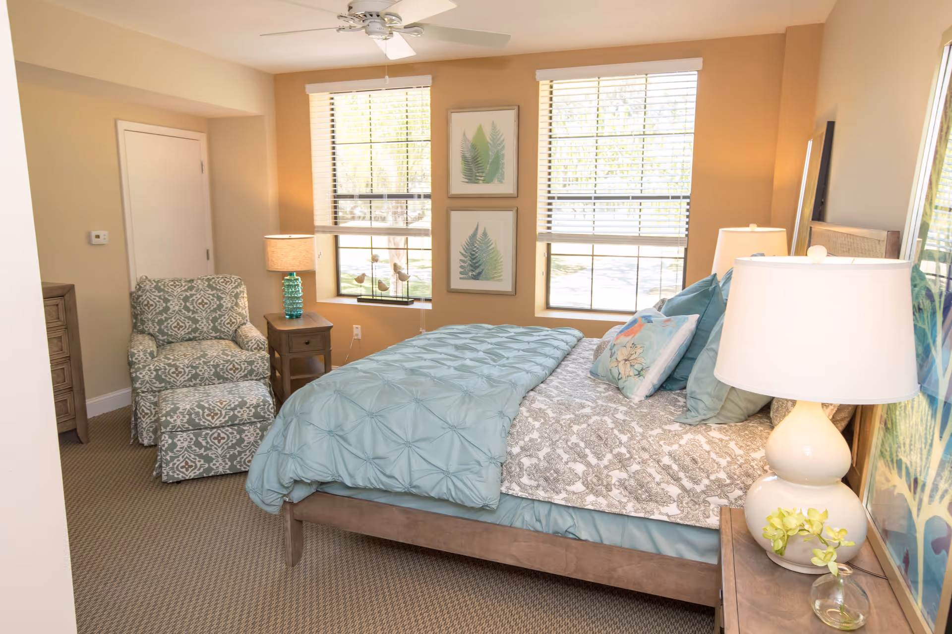 Sunlit, tastefully decorated bedroom with a made bed, bedside lamps, an upholstered armchair, and two windows.