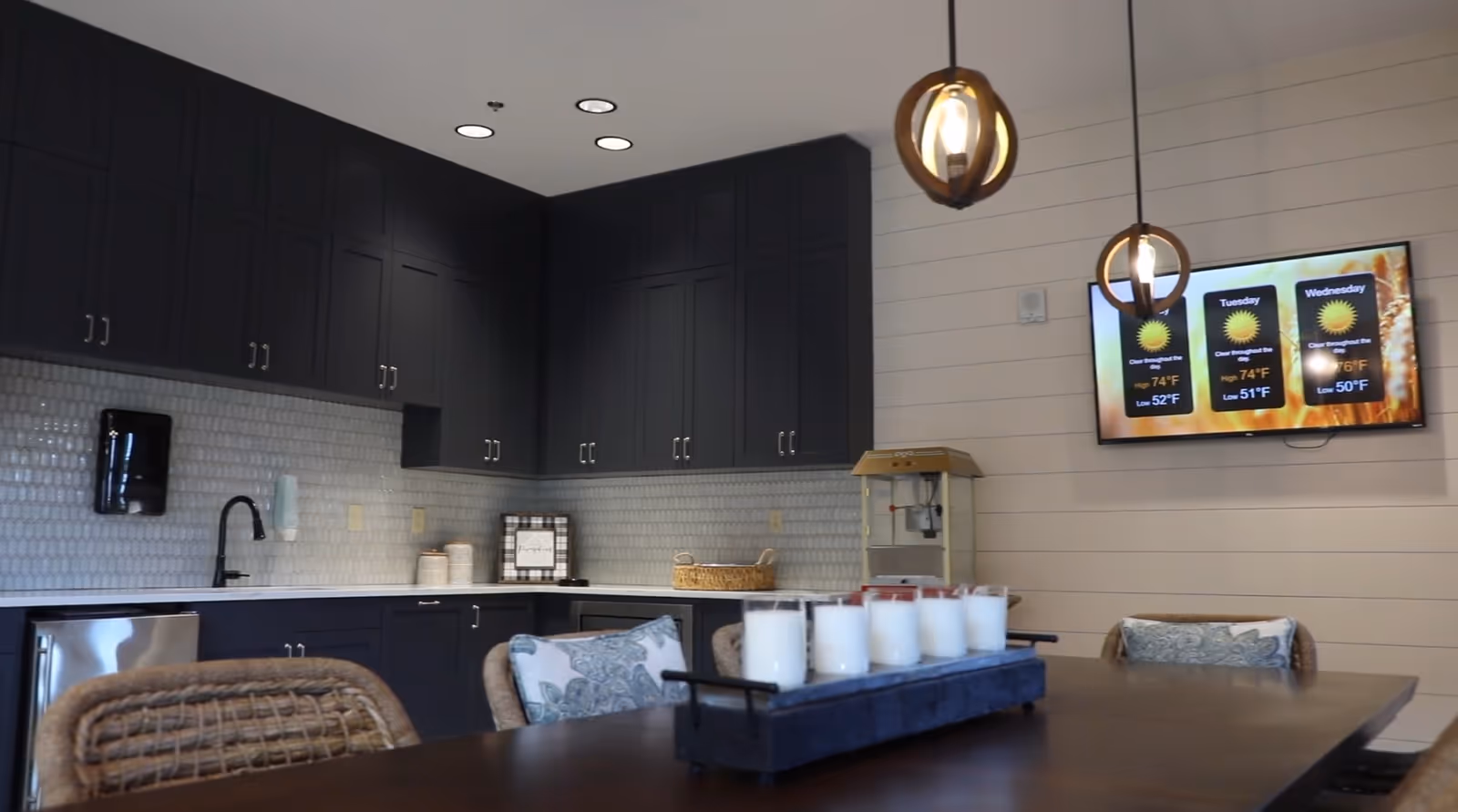Communal kitchen and dining area with dark cabinets, countertop, pendant lights, and a TV displaying the weather.