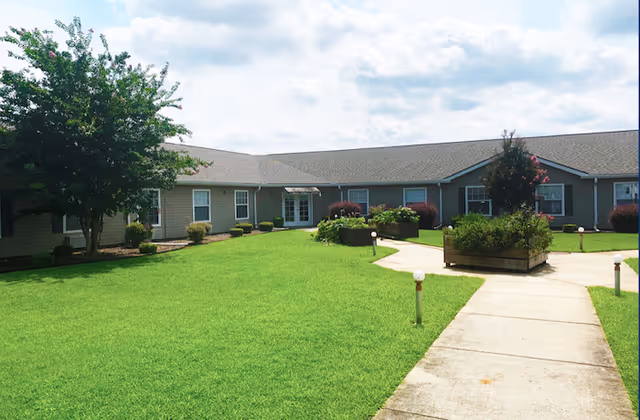 Outdoor view of a single-story building with a gray roof and beige siding, surrounded by a well-maintained green lawn, small bushes, and trees. A concrete walkway leads through the grass to the building entrance under a partly cloudy sky.