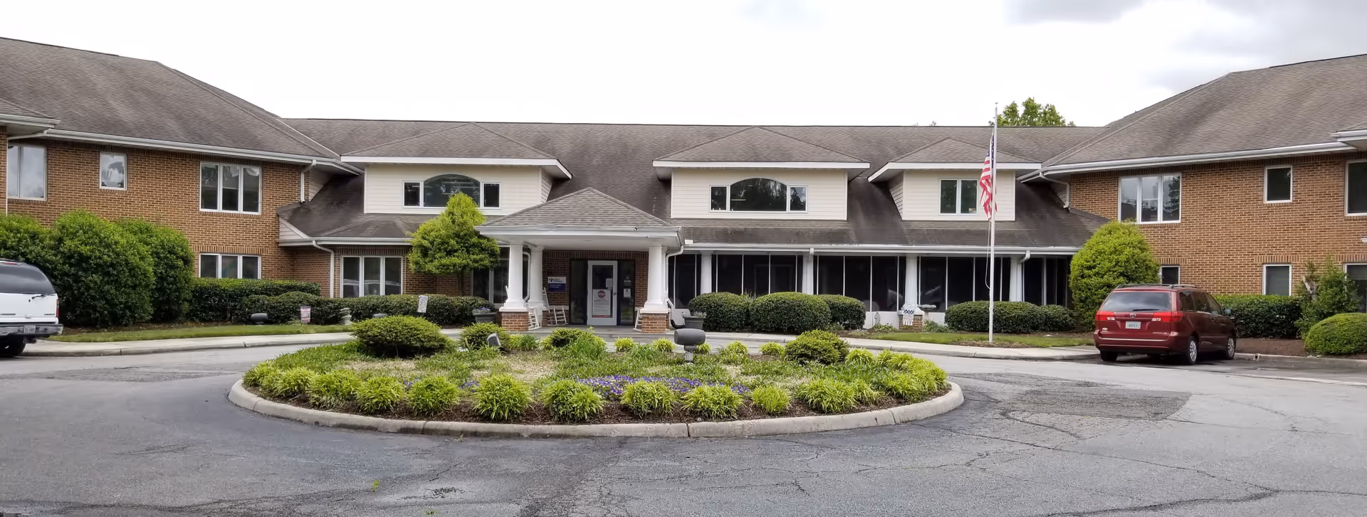Front exterior view of a two-story senior living facility building with a circular driveway and landscaped roundabout with shrubs and flowers. Two vehicles are parked near the entrance, and an American flag is displayed on a flagpole near the building entrance.