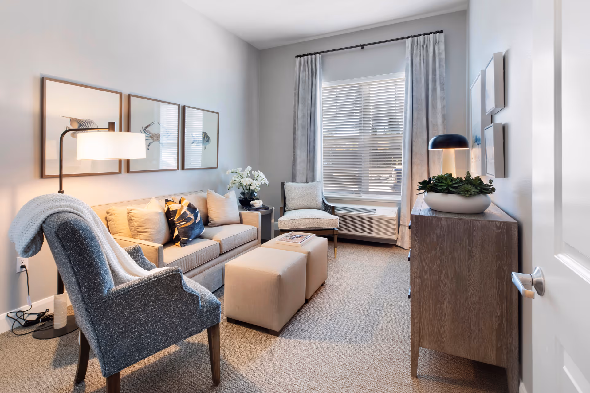 A cozy living room with a beige sofa adorned with pillows, a gray armchair with a blanket draped over it, two beige ottomans, a wooden sideboard with a lamp and a plant, and a window with blinds and curtains letting in natural light.
