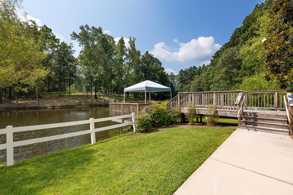 A peaceful outdoor scene at Lakeview Estates - Memory Care Community featuring a wooden deck with stairs leading down to a grassy area beside a calm pond. There is a white canopy tent on the deck, surrounded by green trees and bushes under a partly cloudy blue sky.