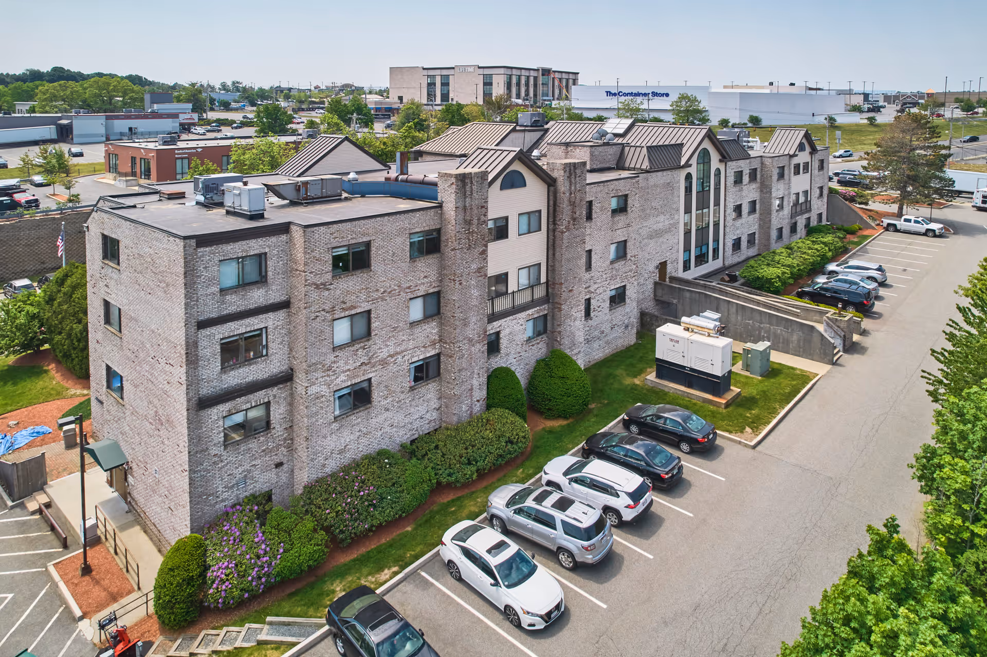 Aerial view of a multi-story brick building with a parking lot in front, surrounded by greenery and other commercial buildings in the background under a clear sky.