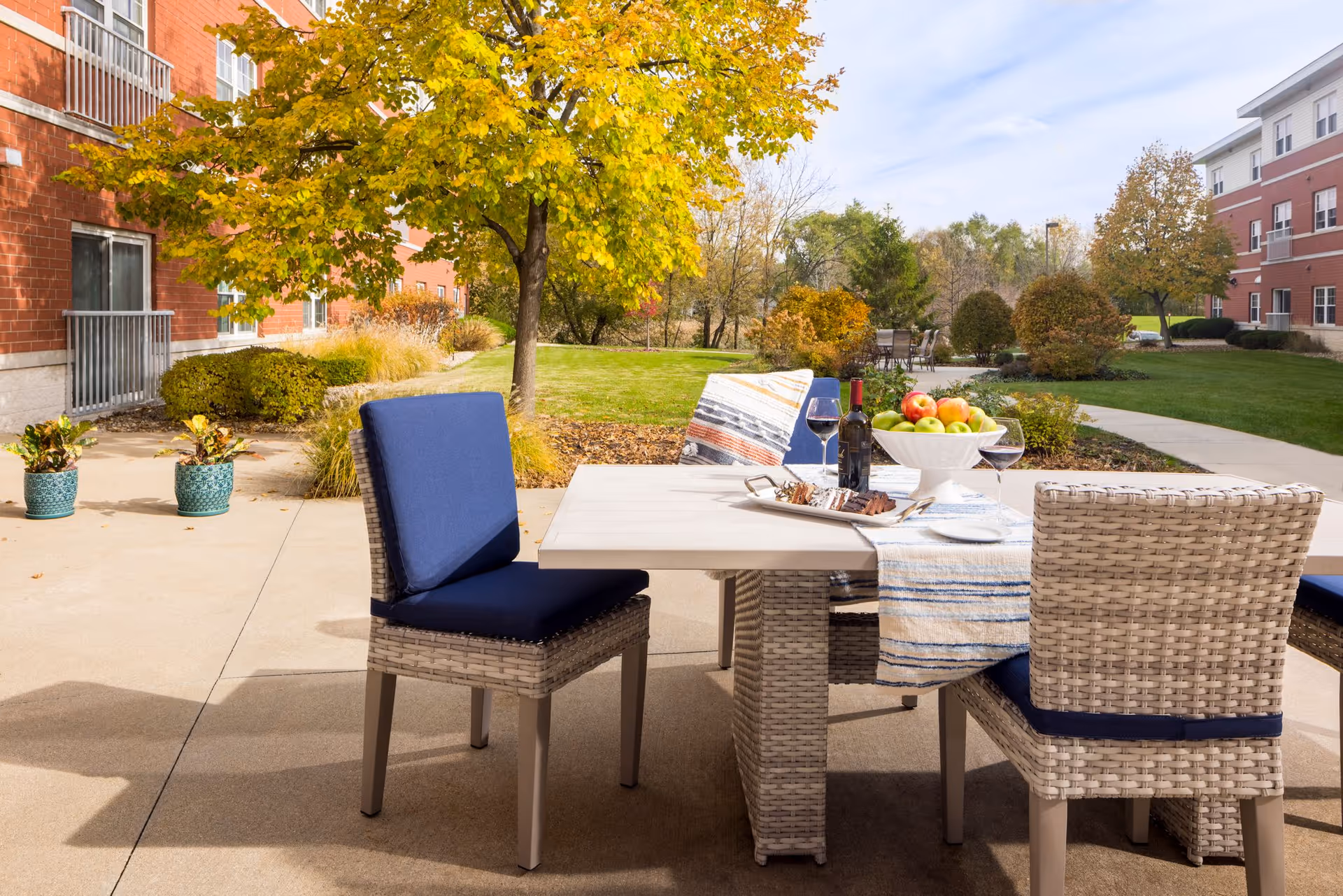 Outdoor patio area at Hanover Place with a table and four wicker chairs with blue cushions. The table is set with a striped table runner, a bowl of apples, a bottle of wine, two glasses of red wine, and a plate of food. Surrounding the patio are green lawns, trees with autumn foliage, and red brick buildings.