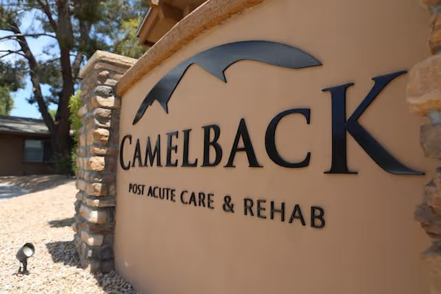 Close-up view of a stone and stucco sign for Camelback Post Acute Care & Rehab with trees and a building in the background.