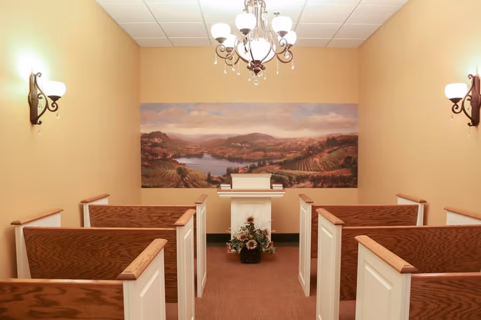 Small chapel room with wooden pews arranged in two rows facing a white podium adorned with a floral arrangement. The walls are painted beige and feature a large landscape painting of a countryside with hills and a river. The room is softly lit by wall sconces and a chandelier hanging from the ceiling.