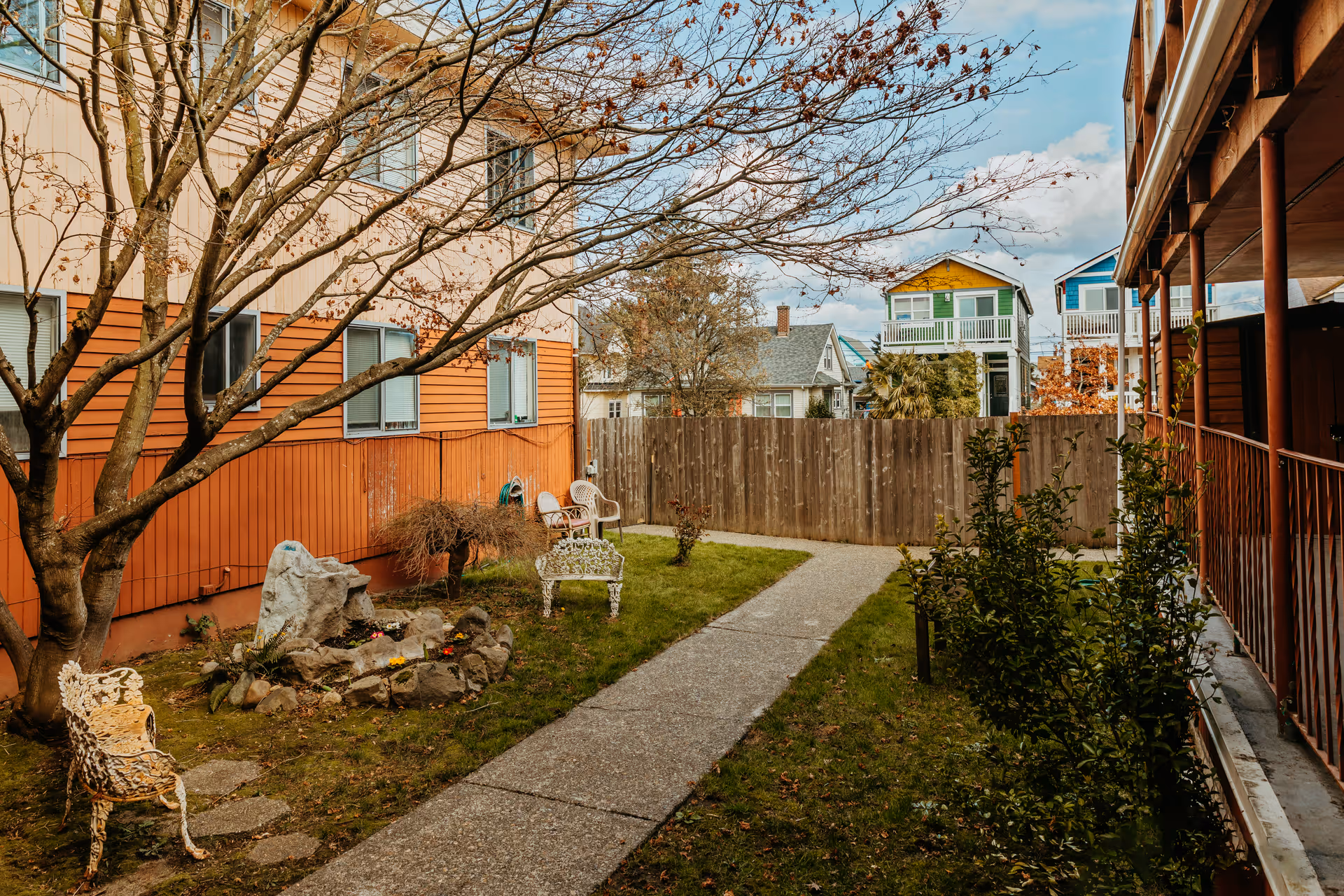 Outdoor courtyard area at 6th Avenue Senior Living with a concrete pathway, grass, a leafless tree, garden chairs, and a small rock garden. Surrounding buildings and a wooden fence are visible in the background under a partly cloudy sky.