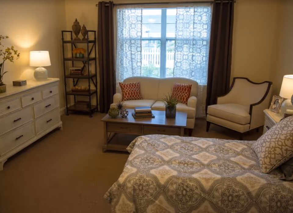 A cozy bedroom in a senior living facility featuring a bed with patterned bedding in the foreground, a white loveseat with two red patterned pillows in front of a window with lace curtains, a beige armchair, a wooden coffee table with decorative plants and books, a white dresser with a lamp and decorative items, and a black metal shelving unit with various decor pieces.