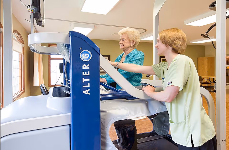 An elderly woman using an AlterG anti-gravity treadmill assisted by a healthcare worker in a light green uniform inside a well-lit room with large windows.