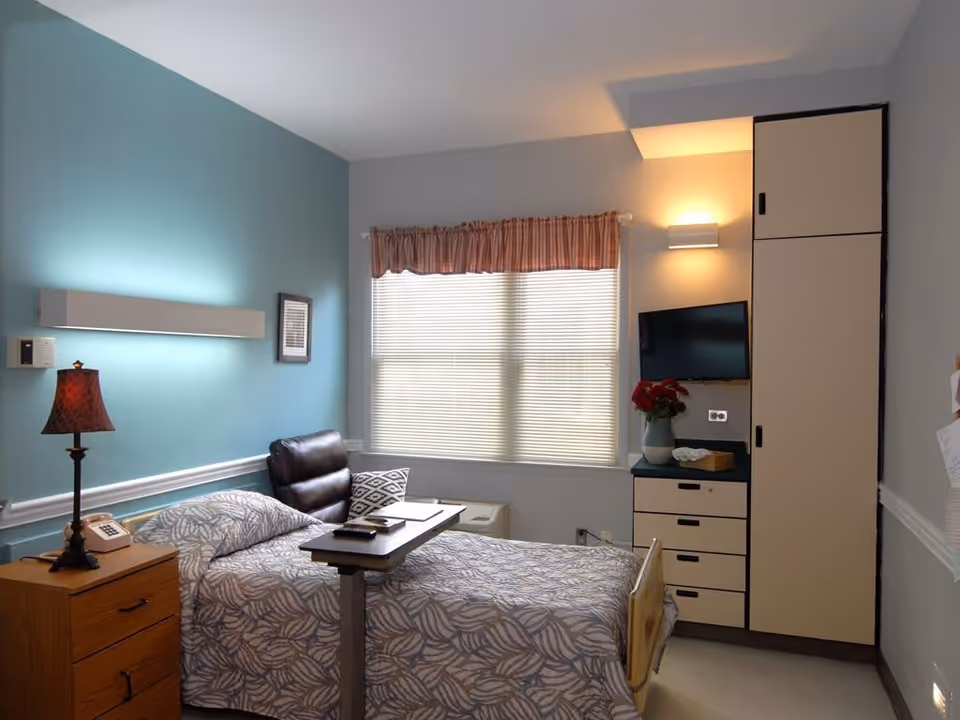A single bed in a senior living facility room with patterned bedding, a bedside table with a lamp and telephone, a small overbed table, a black leather chair, a window with blinds and a pink valance, a mounted flat-screen TV, and built-in cabinets with a vase of red flowers on the counter.