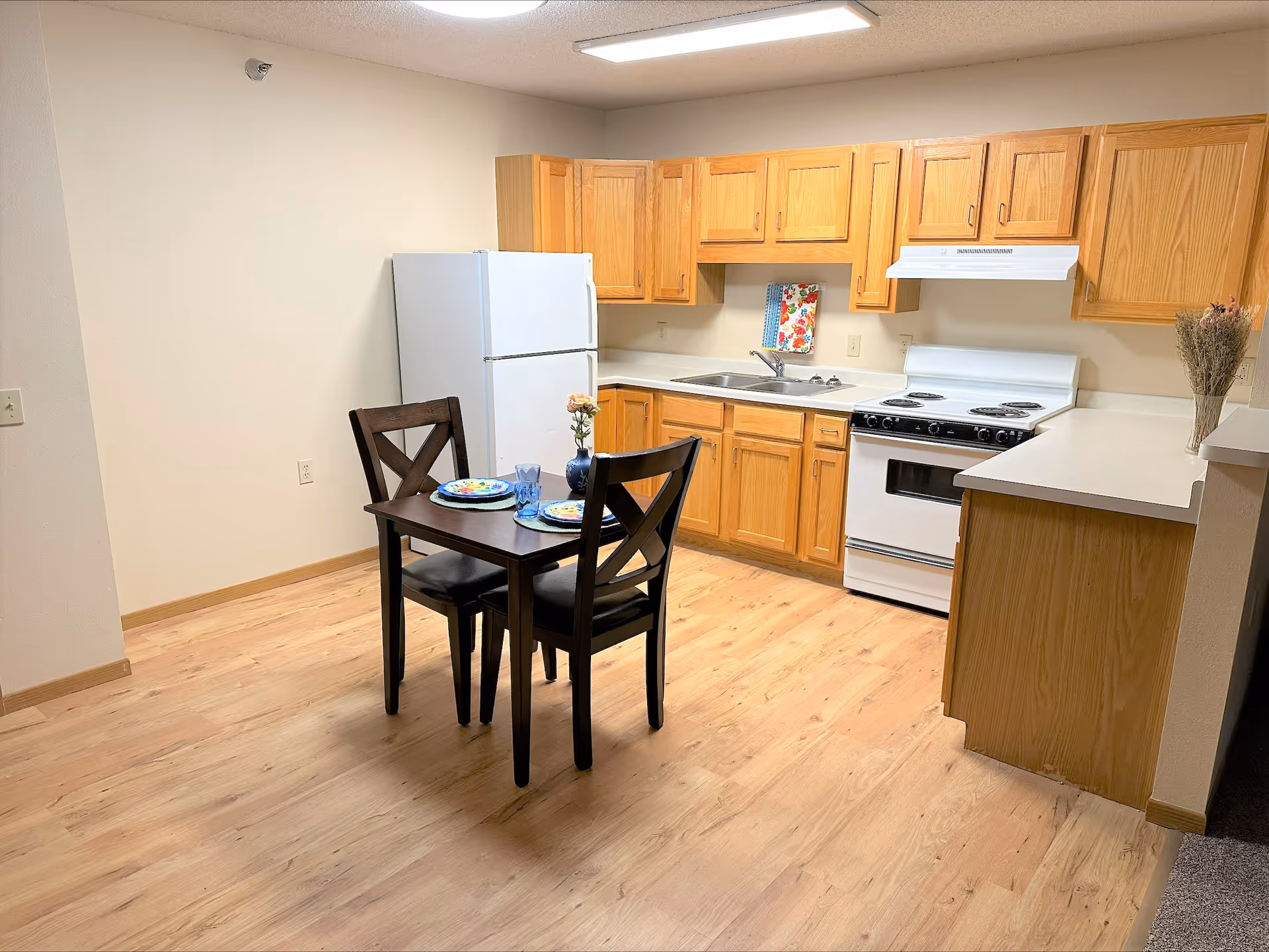 A small kitchen area with wooden cabinets, a white refrigerator, a white stove with an oven, and a sink. In front of the kitchen is a dark wooden dining table set with two chairs, two plates, two glasses, and a small vase with a flower. The floor is light wood, and the walls are painted beige.