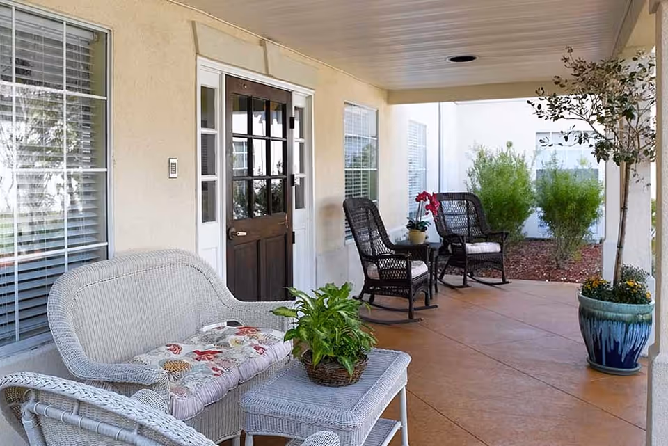 Covered front porch with wicker loveseat, table, rocking chairs, potted plants, and a dark wooden entry door.