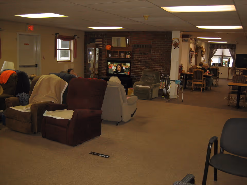 A common area in a senior living facility with several recliner chairs arranged facing a television mounted on a brick wall. Some elderly residents are seated in the chairs watching TV. In the background, there are tables and chairs where other residents are seated, and a walker is visible near a pillar. The room is carpeted and has fluorescent ceiling lights.